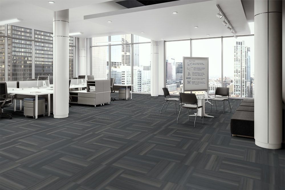 Fiction flooring in the open office. At one side are occupied work desks with attached credenzas. A portable white board is placed to the side of one round cafe table, with instructions written upon it. The full height windows look out to the city.