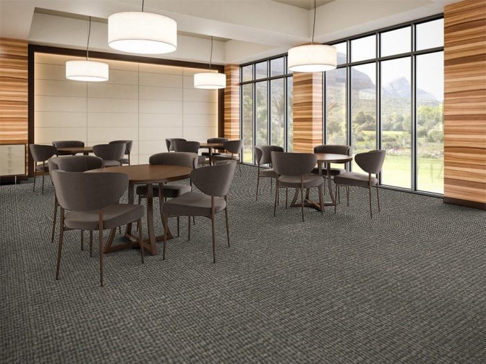 Structure flooring in a dining area. Matching round tables are placed towards one end, with 4 cushioned chairs placed at each. Sunlight comes in from the tall windows.