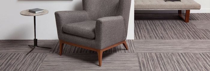 Studio photography, looking into a room lined with Timber carpeting. A round cafe table and plush chair has been placed against the wall, with a long cushioned bench against the wall behind.