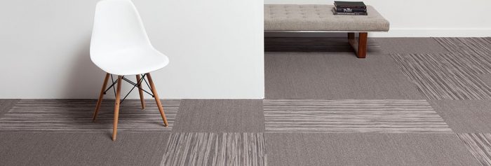 Studio photography, looking into a room lined with solid colored carpeting, with Timber planks breaking up the design. A white plastic chair has been placed against the wall, with a long cushioned bench against the wall behind.