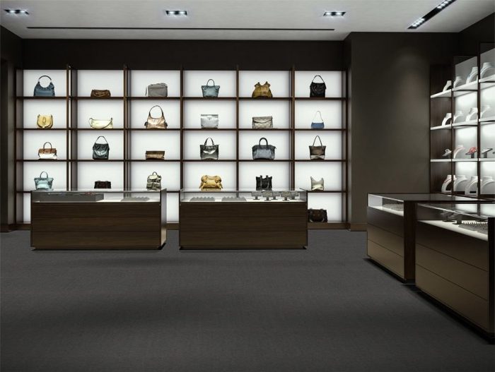 Approaching a counter in an accessories store. The floor uses Transit model carpeting, with the Pass color scheme. Behind the two counters are shelves which hold designer purses.