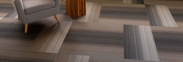 Studio shot of Umbra II carpeting in a grey herringbone pattern. On the left is a wooden cubic end table, with a cushioned chair beside.