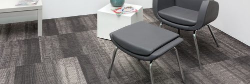 Studio shot of Well Versed model carpeting. The corner of this room contains a plush upholstered chair with matching ottoman. At the side is a white cube end table and a long table placed against the wall.