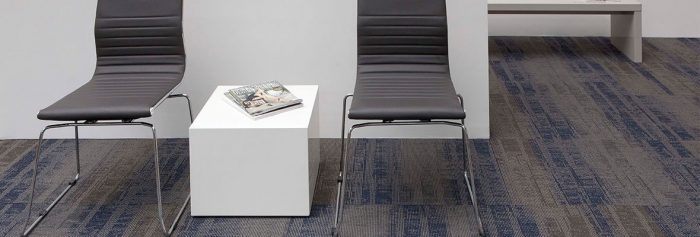 Studio photography, looking into a room lined with Well Versed model carpet squares. Two chairs and cube table have been placed against the wall, with a long cushioned bench against the wall behind.