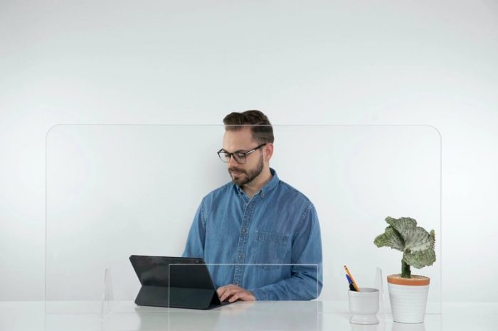 A man sits at a large table, working on his computer. A Counter Shield screen separates him from the space in front.